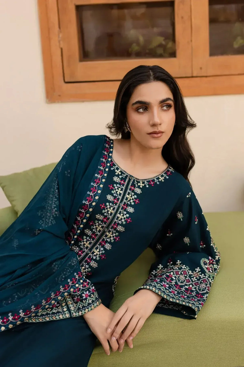 Woman in teal embroidered traditional kurta sitting on light green couch with wooden window in background, Sitarah Clothing